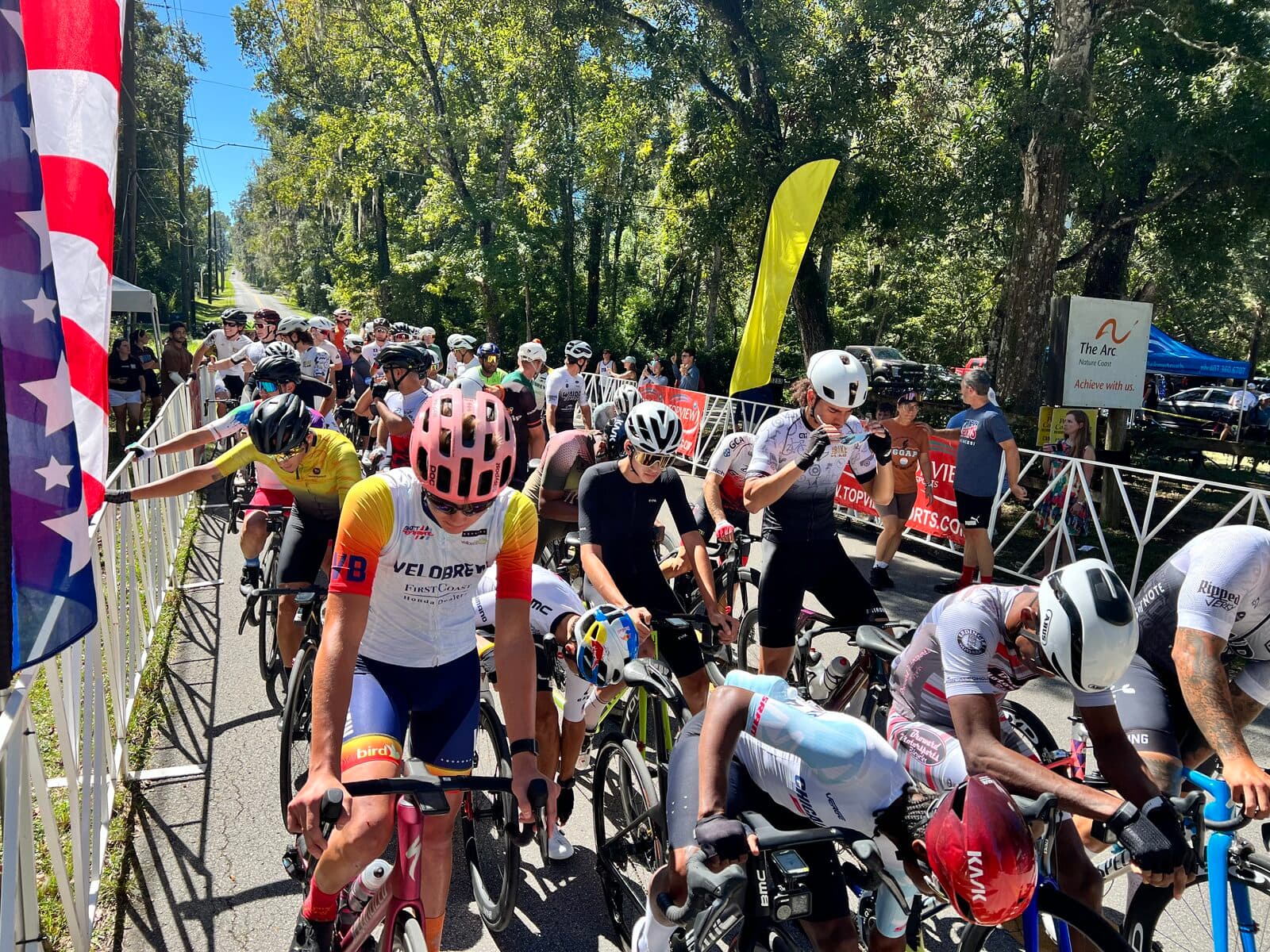 Olen Lokkesmoe at a criterium race start line surrounded by competitors, wearing Velobrew cycling kit with pink helmet